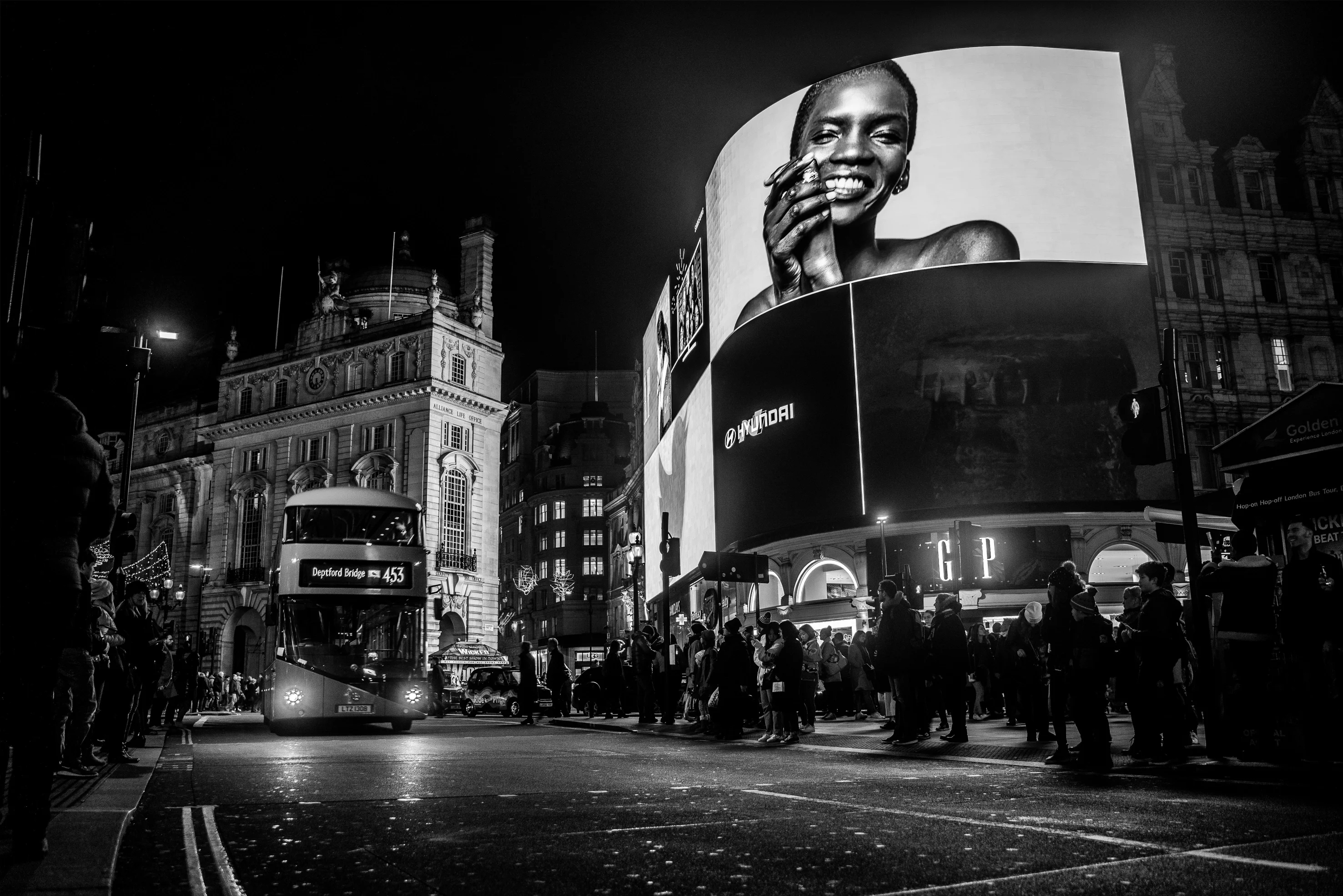 Svart och vit bild på en gata där man i vänstra hörnet ser en klassisk dubbeldäckare i London. I högra delen av bilden ser man en stor bildbord med en kvinna som håller händerna i ansiktet och ler stort. Under bilboarden är det en massa människor.    