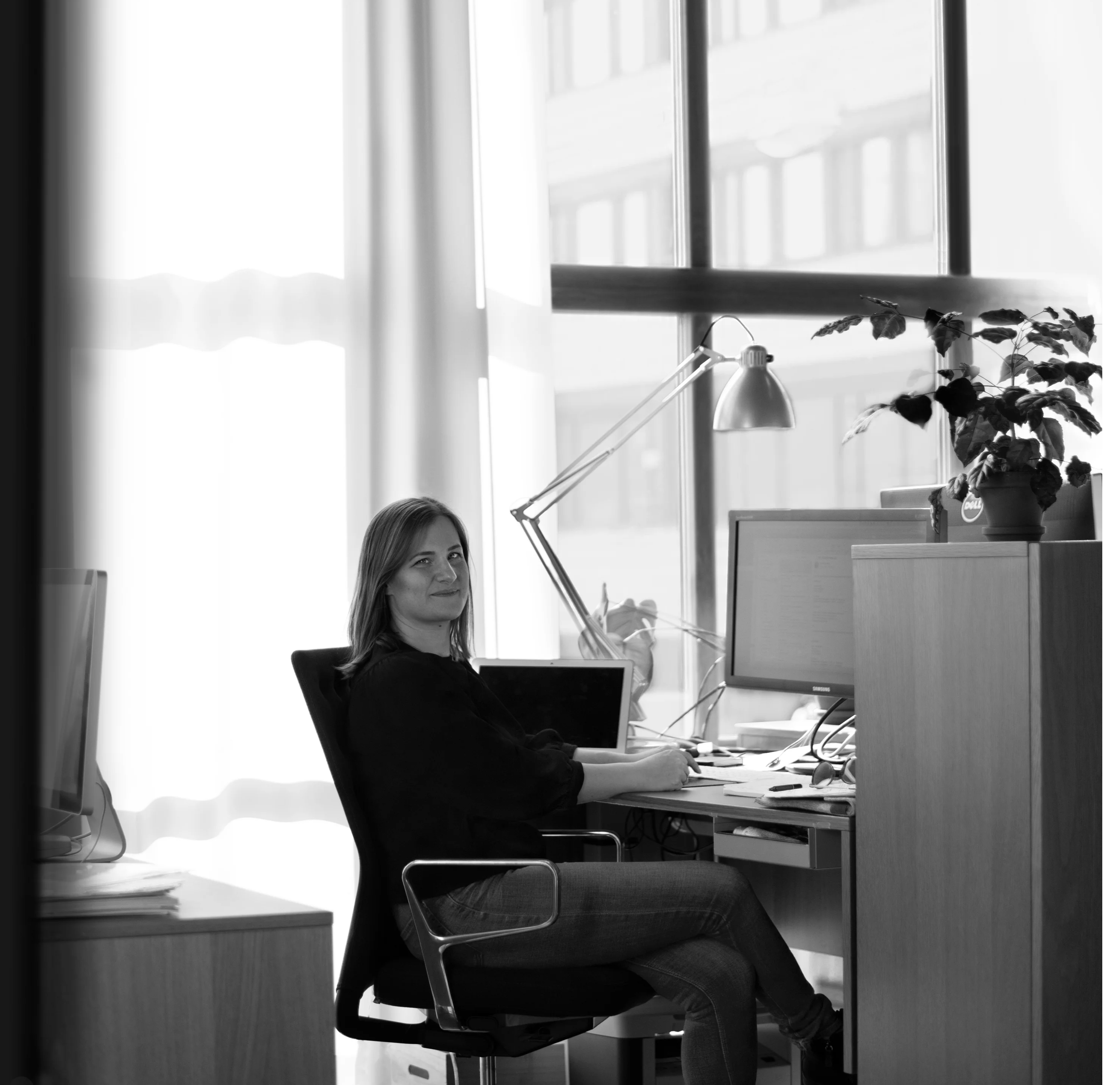 Svartvit bild, kvinna sitter till vänster om hennes skrivbord, höger axel mot kameran, huvudet vändt mot kameran hon ler. Händerna på skrivbordet framför dator, sidleds till datorn en bordslampa. Bakgrunden stort fönster med genomskinliga gardiner. 
