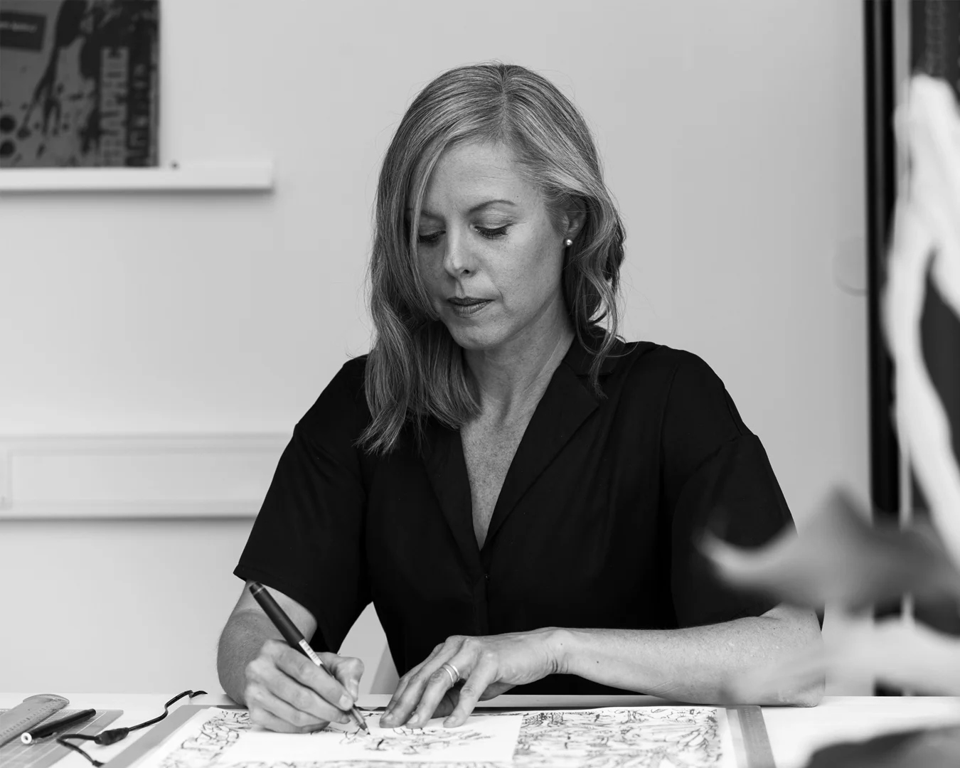Svart och vit bild, ljushårig kvinna centrerad med huvudet blickande nedåt, håller i en penna och skissar på ett papper -NyStudio